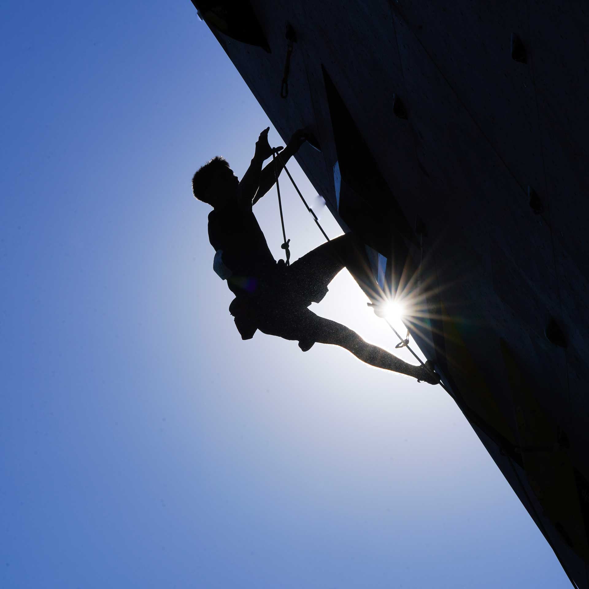 A lead climbing competitor reaches to clip a draw, with the sun in the background